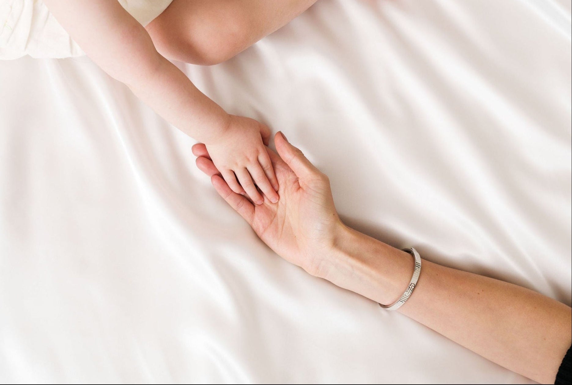 Mother holding baby's hand on silky sheets, wearing her Vivi Coo silver personalised bracelet.